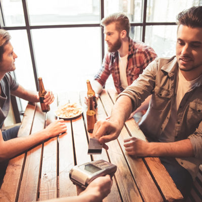 Handsome friends are drinking beer talking and smiling while resting at the pub. One guy is paying using a credit card
